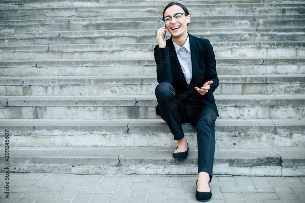 Cheerful Attractive business woman in a suit and glasses speaks on a smart phone sitting on gray concrete stairs. Cheerful Attractive business woman in a suit and glasses speaks on a smart phone sitting on gray concrete stairs.