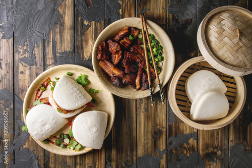 Asian sandwich steamed gua bao buns with pork belly, greens and vegetables served in ceramic plate over dark wooden plank background. Asian style fast food dinner. Flat lay, space