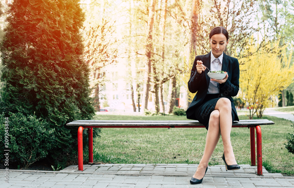 Healthy nutrition on break. Modern smiling business woman in a black suit with a fork eating a vegetable salad outdoors Healthy nutrition on break. Modern smiling business woman in a black suit with a fork eating a vegetable salad outdoors