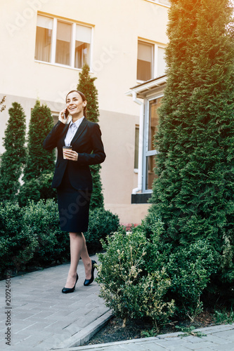 Working business conversation. A modern smiling business woman in a black suit is walking down the street and talking on the phone with a client