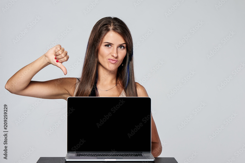 Discontent woman showing blank black laptop computer screen, looking at camera and gesturing thumb down Discontent woman showing blank black laptop computer screen, looking at camera and gesturing thumb down