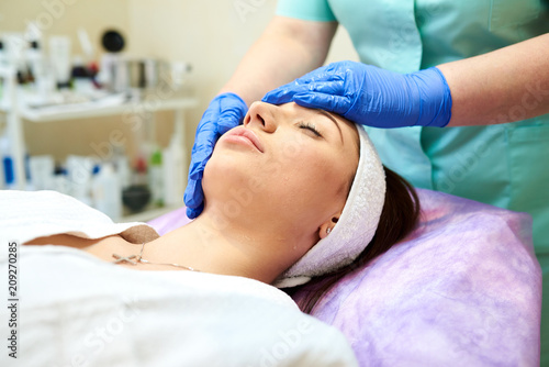 Cosmetologist masseuse cleaning woman face at spa in cosmetic cabinet under a lamp. Clean fresh skin, wearing white bathrobe and hair bandage, doing cosmetic procedure. Facial peeling mask applying.