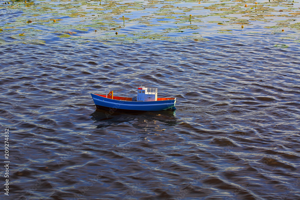Naklejka premium Cute radio controlled blue toy boat on water surface. Dark lake water and water lilies on background. Hobby concept.