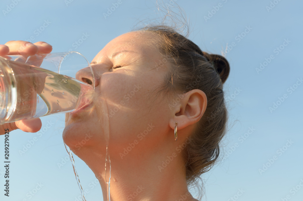 A simple European young woman is drinking water from a glass beaker against a blue sky. - obrazy, fototapety, plakaty 