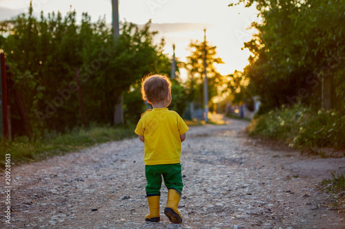a little boy walks along the country road in rubber yellow boots and a bright T-shirt.