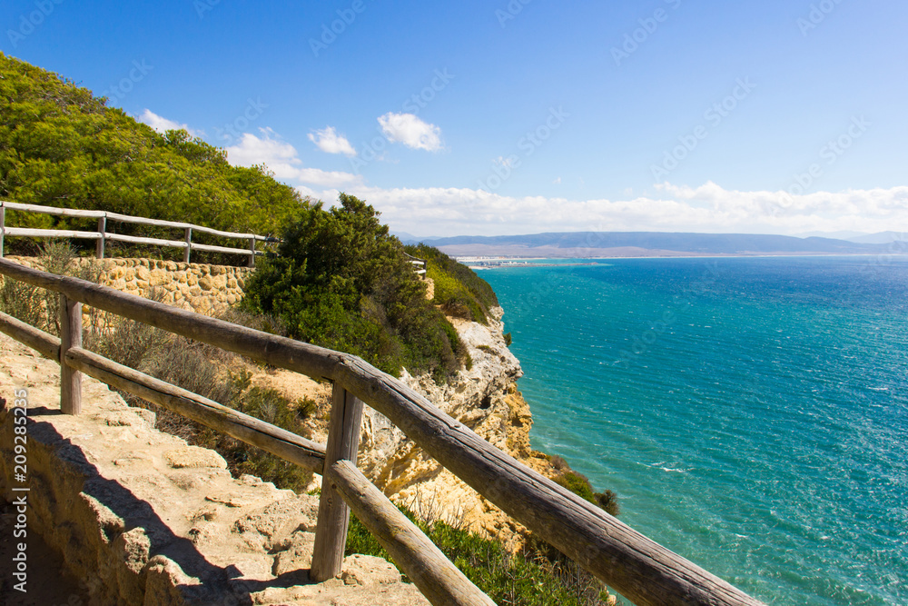 Wood railing along walk path of nature park by the sea with Barbate town on the background in Cadiz, Andalusia. Hiking, trek adventure, coastline landscape concept