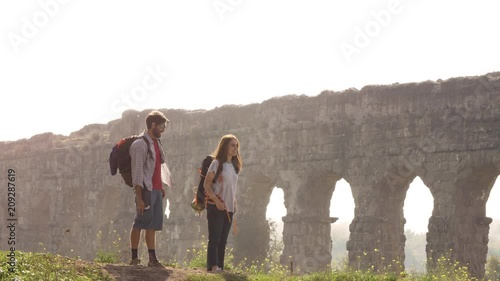 Young lovely couple backpackers tourists reading map pointing directions roman aqueduct arches in parco degli acquedotti park ruins in rome on romantic misty sunrise with guitar and sleeping bag slow photo