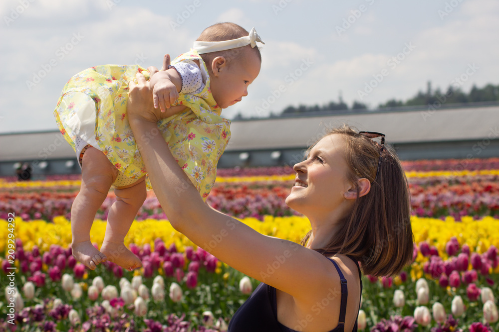 Young New Mom with Baby Infant in Fancy Dress in Tulip Flower Field Young New Mom with Baby Infant in Fancy Dress in Tulip Flower Field
