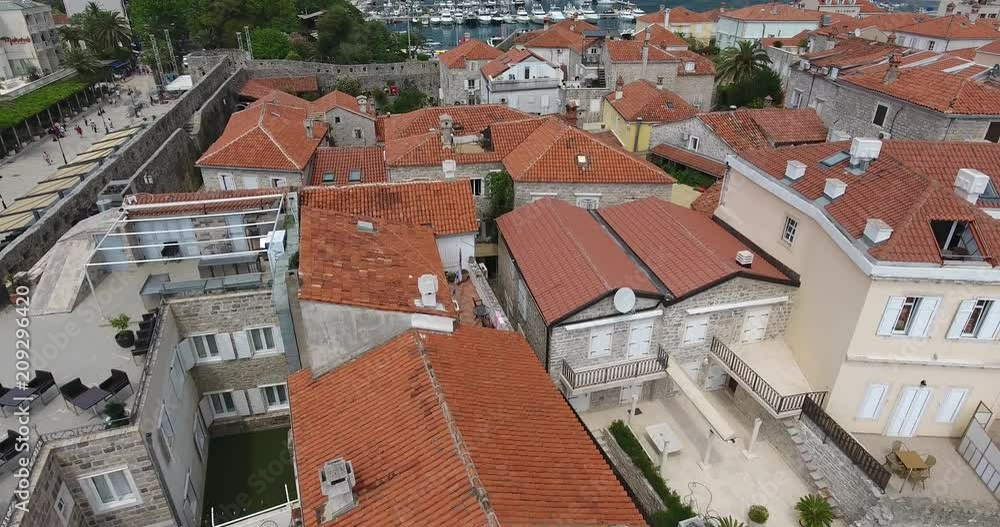 Flight above the Old Town of Budva, Montenegro. Aerial view of Old Town Budva, red roofs of its stone houses in the traditional Balkan style, ancient church and a seashore with boats Flight above the Old Town of Budva, Montenegro. Aerial view of Old Town Budva, red roofs of its stone houses in the traditional Balkan style, ancient church and a seashore with boats