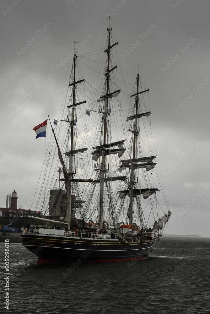 A three-masted modern luxury clipper with historical nineteenth century styling sailing in the North Sea Canal's mouth under the cloudy gray sky, IJmuiden, the Netherlands.