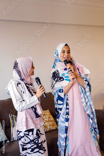 Two young Muslim Malay women unwind and relax as they sing songs on their Karaoke machine at home during the day during Raya. 