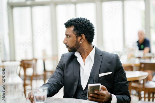 An Indian Asian businessman takes a break from work as he sits in a cafe fiddling through his smartphone. He is well-dressed in a dark suit.