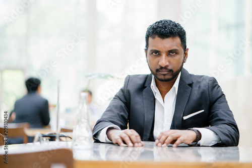 Portrait of a young Indian Asian businessman looking intense and serious at the camera. He is wearing a gray suit and is seated at a table in their office during the day.