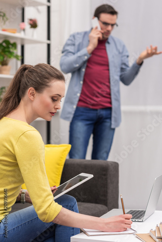 adult woman working with tablet while her boyfriend talking by phone at home