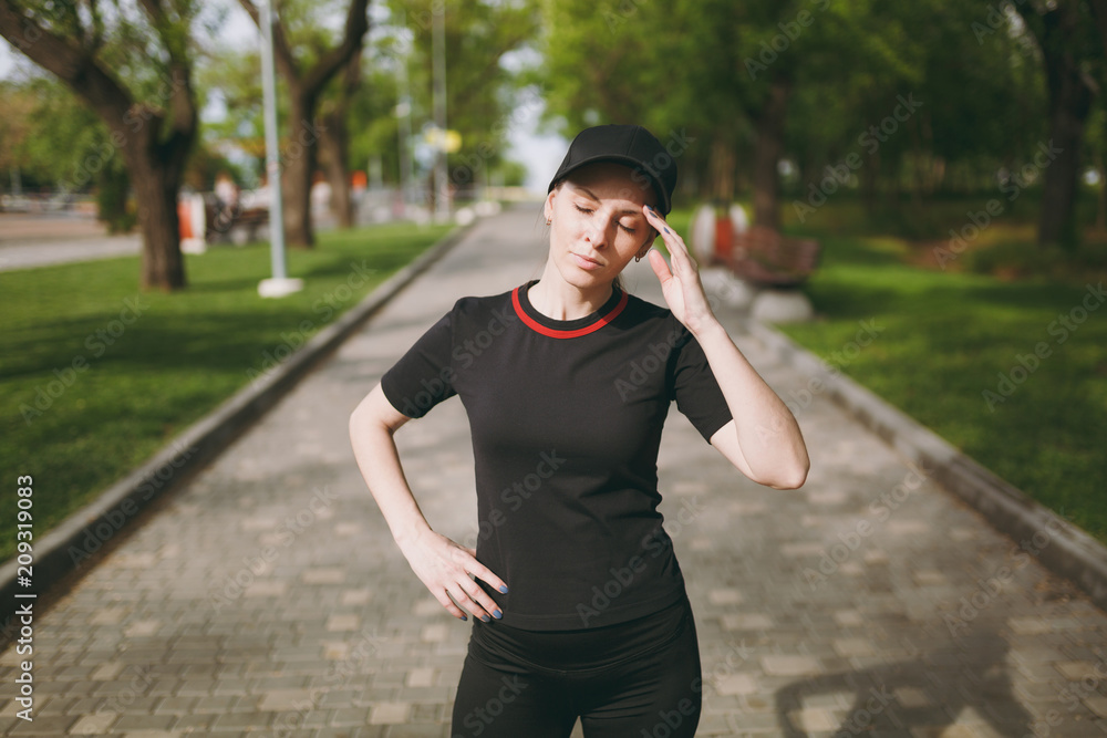 Young tired athletic beautiful brunette girl in black uniform and cap standing, resting and keeping hand near head after running, training in city park outdoors. Fitness, healthy lifestyle concept. Young tired athletic beautiful brunette girl in black uniform and cap standing, resting and keeping hand near head after running, training in city park outdoors. Fitness, healthy lifestyle concept.