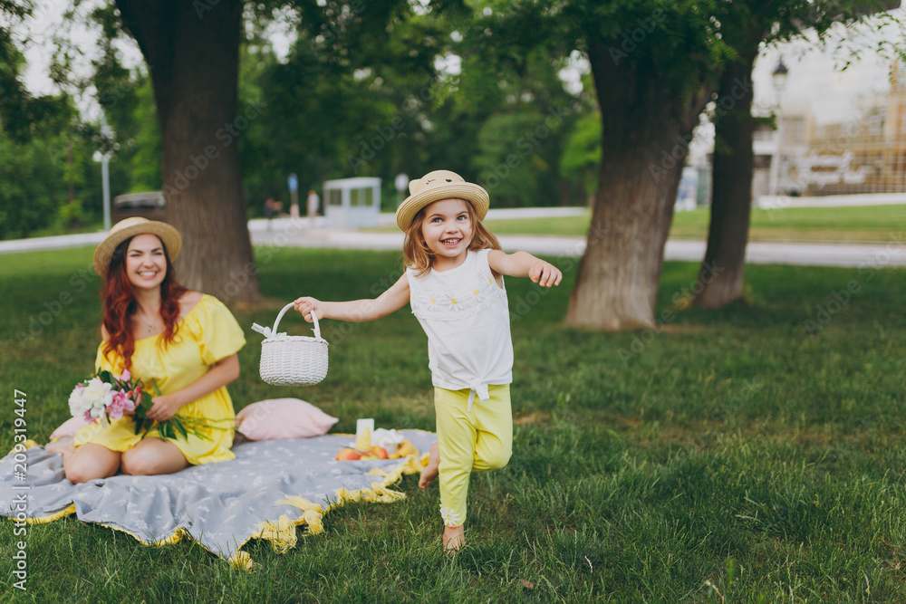 Fototapeta premium Little cute child baby girl with basket play and have fun, rest with woman in yellow clothes on green grass in park. Mother, little kid daughter. Mother's Day, love family, parenthood, childhood.