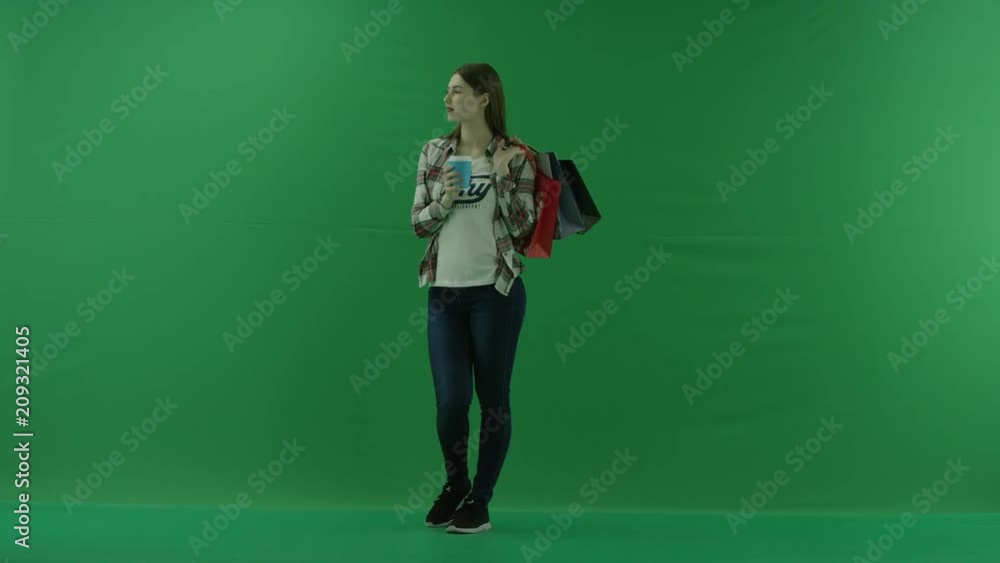 the girl stands in a half-turn, looks at the left, with bags on her shoulder and drinks something from a cup on green screen the girl stands in a half-turn, looks at the left, with bags on her shoulder and drinks something from a cup on green screen