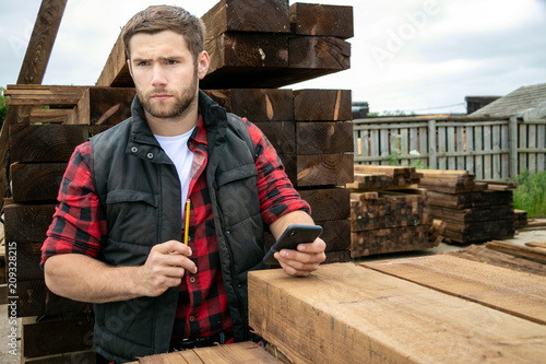 Lumber yard worker, carpenter at wood yard counts inventory with mobile device