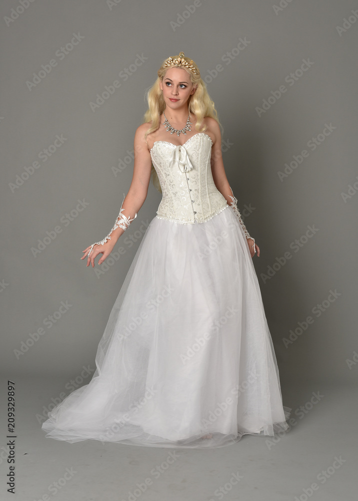 full length portrait of blonde girl wearing white corset gown. standing pose on grey studio background.