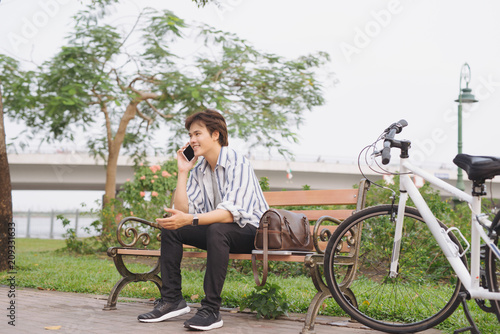 Young man using phone while sitting on bench with bicycle in park