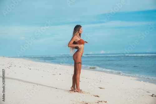 Side view of a young beautiful slender girl with a flat belly and a round booty admiring the blue ocean standing on the shore of a white sandy beach. The concept of recreation, travel, tourism.