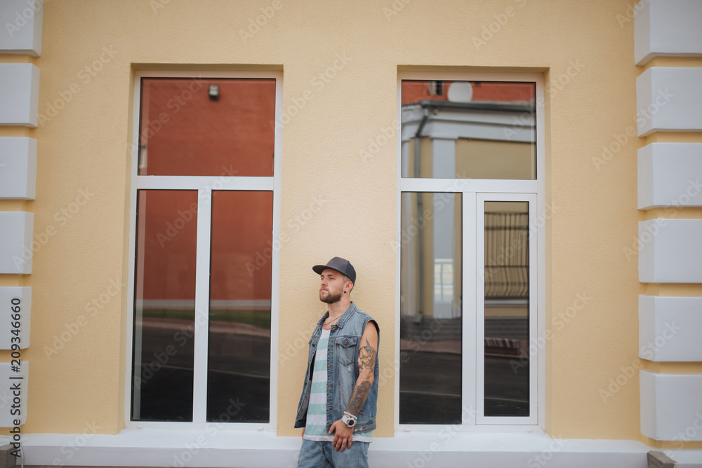 Enjoying casual style. Hipster and urban fashion. Lifestyle portrait concept. Attractive guy in a cap next to wall. Man in a cap with tattoos standing between the windows another view