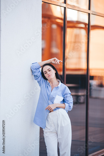 A beautiful woman in a shirt and pants is standing by the mirror window of the balcony of the building. 