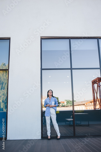 A beautiful woman in a shirt and pants is standing by the mirror window of the balcony of the building. 