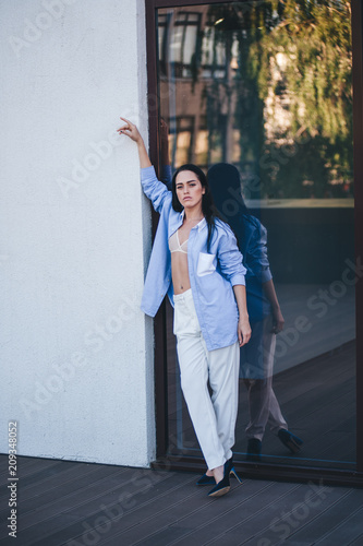 A beautiful woman in a shirt and pants is standing by the mirror window of the balcony of the building. 