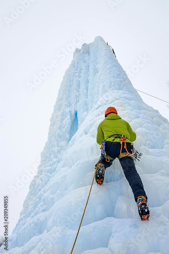 Alpinist man with ice tools axe in orange helmet climbing a large wall of ice. Outdoor Sports Portrait