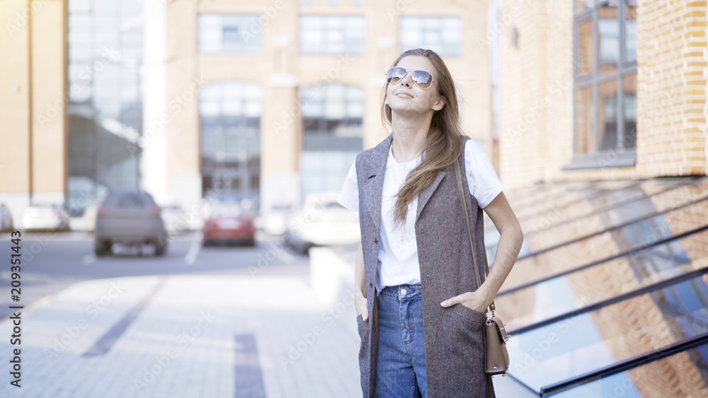 A young smiling girl long hair dressed in a jacket and jeans is wearing sunglasses outside hands in the pockets