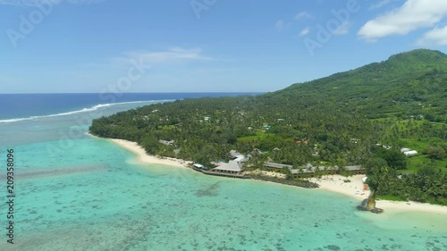 AERIAL: Flying near large luxury resort set right on the beautiful white sand beach. Breathtaking shot of remote island hotel with a perfect view of the lush mountains and turquoise Pacific Ocean.