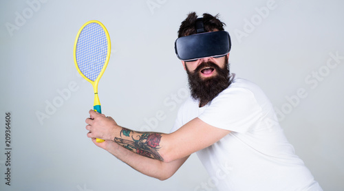 Bearded man doing sports in virtual reality. Hipster in VR headset with  tennis bat isolated on gray background. Man with bushy beard playing  computer simulated tennis game, online gaming concept foto de