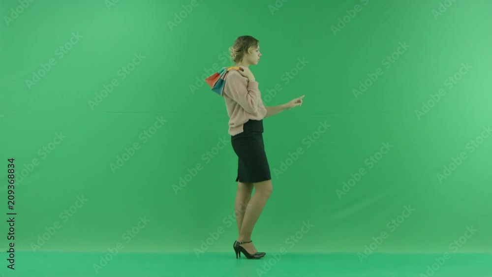 Pretty shopping woman stands in front of the digital screen with shopping bags and touching the screen. Female shopper holding shopping bags sideways on green background in studio. Pretty shopping woman stands in front of the digital screen with shopping bags and touching the screen. Female shopper holding shopping bags sideways on green background in studio.