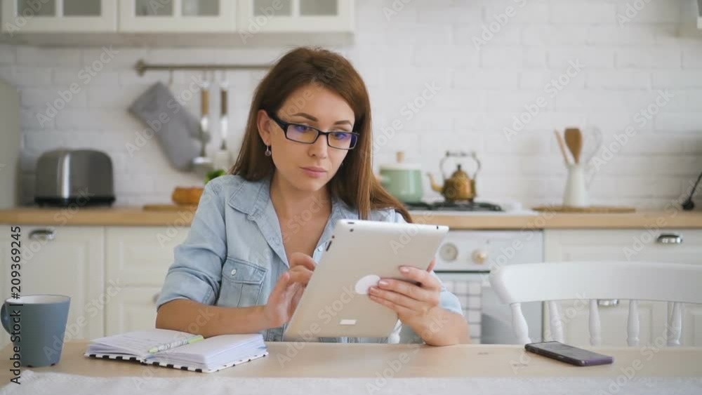 Young businesswoman is using tablet sitting at table in home kitchen, beautiful lady in jeans clothes working at desk with device and notebook in modern interior. Concept: business person, social Young businesswoman is using tablet sitting at table in home kitchen, beautiful lady in jeans clothes working at desk with device and notebook in modern interior. Concept: business person, social