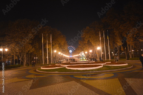 background of the Ferris wheel along the promenade are benches  trees and yellow lanterns and a round flower square