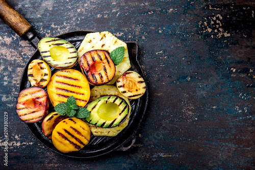 GRILLED FRUITS. Grill fruits - pineapple, peaches, plums, avocado, pear on black cast iron grill pan. Dark background. Top view. Copy space photo