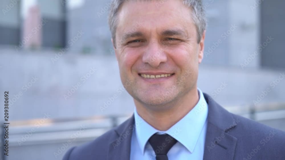A gray-haired middle-aged man in a suit looks at the camera against the backdrop of a business center. Business and success. portrait to the waist 4K A gray-haired middle-aged man in a suit looks at the camera against the backdrop of a business center. Business and success. portrait to the waist 4K