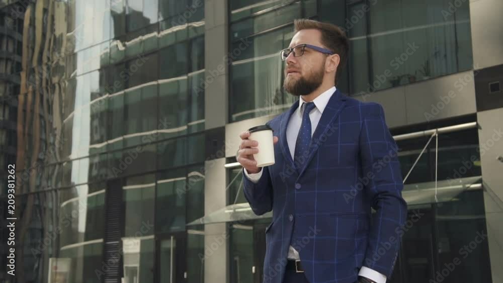 Confident businessman is drinking drink, looking at watch, standing near office building, bearded guy wearing stylish suit is having coffee break, resting during working day in summer day. Concept Confident businessman is drinking drink, looking at watch, standing near office building, bearded guy wearing stylish suit is having coffee break, resting during working day in summer day. Concept