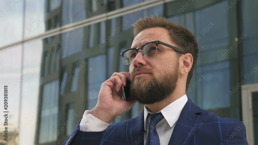 Portrait of solid young man talking on phone standing on modern building backgroung, bearded businessman wearing glasses is makes call, discussing startup project on street in summer day. Concept Portrait of solid young man talking on phone standing on modern building backgroung, bearded businessman wearing glasses is makes call, discussing startup project on street in summer day. Concept