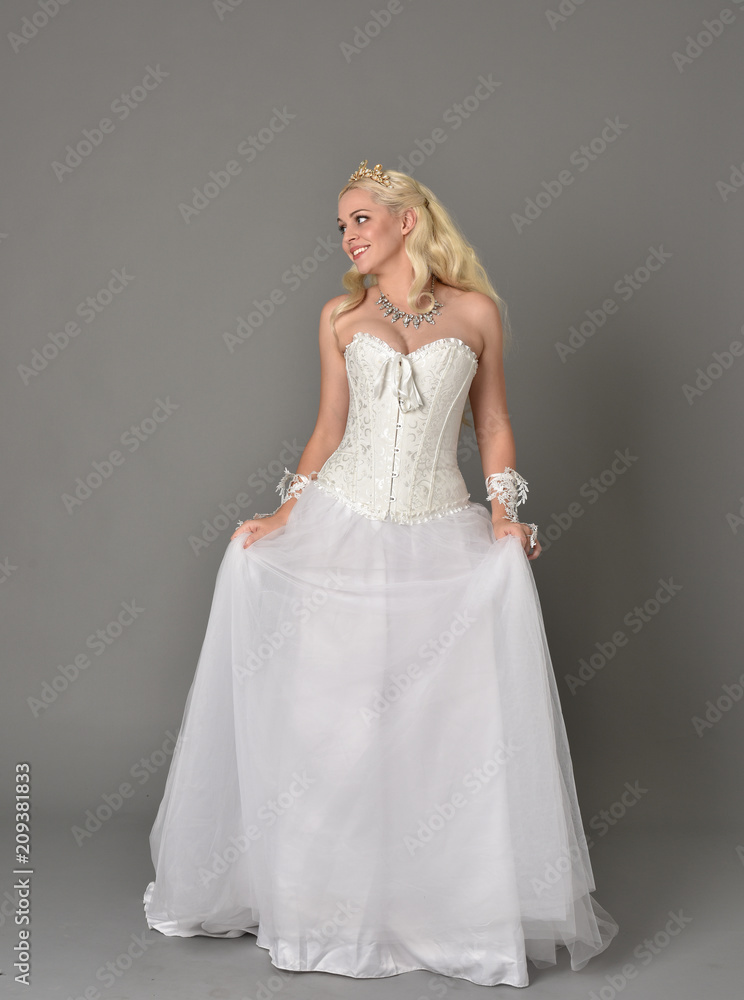 full length portrait of blonde girl wearing white corset gown. standing pose on grey studio background.