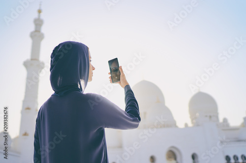 Traveling by Unated Arabic Emirates. Pretty young woman in traditional abaya taking photo with smartphone in the Sheikh Zayed Grand Mosque, famous Abu Dhabi sightseeing. photo