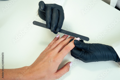 close-up hands of manicurist professional (master of manicure) woman in a black rubber gloves is apply a nail file of client in a beauty spa salon.