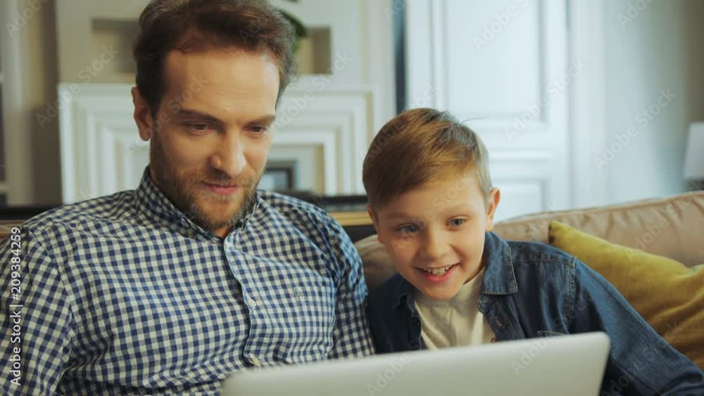 Handsome man with a beard sitting together with his little son on the couch and doing something on the laptop computer in the living room. Portrait shot. Close up. Indoor Handsome man with a beard sitting together with his little son on the couch and doing something on the laptop computer in the living room. Portrait shot. Close up. Indoor