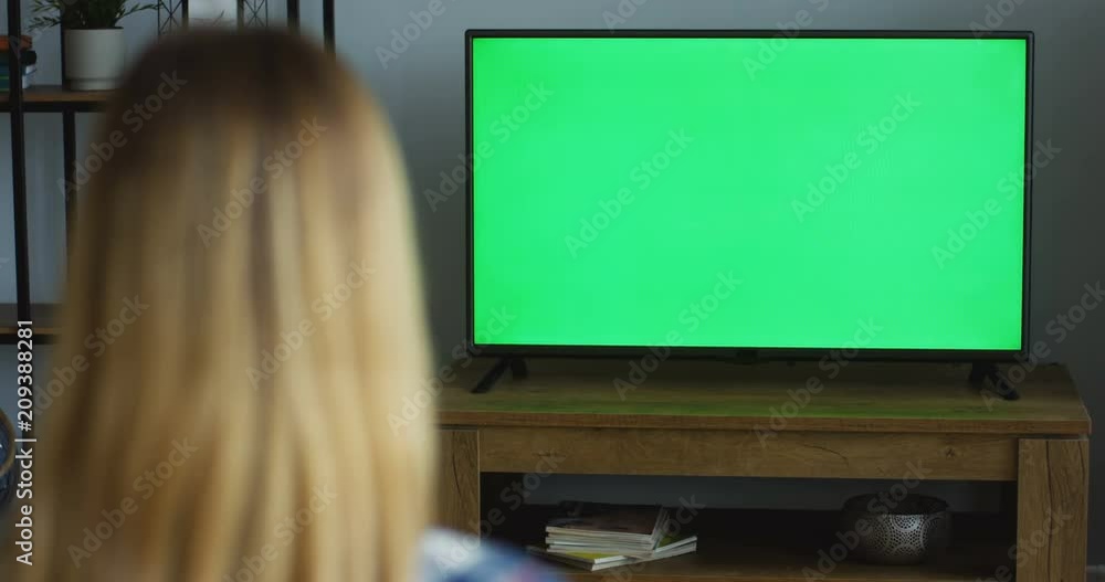 View from the back on the blonde woman sitting in the living room and watching TV with green screen. Chroma key. Indoor View from the back on the blonde woman sitting in the living room and watching TV with green screen. Chroma key. Indoor