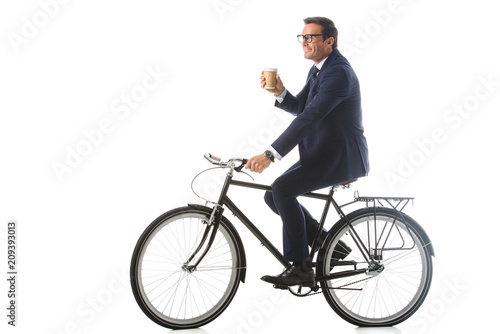 smiling businessman in eyeglasses riding bicycle and holding paper cup of coffee isolated on white background