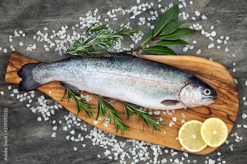 Rainbow trout healthy heart food on an olive wood board, with rosemary and bay leaf herbs, course sea salt and lemon on marble background. High in omega 3 fatty acid.
