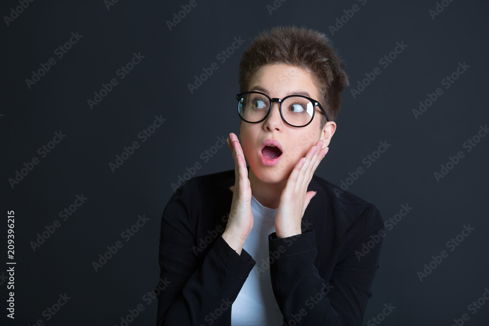 beautiful young girl with short hair, in a black suit and wearing glasses on a black background with a frightened expression beautiful young girl with short hair, in a black suit and wearing glasses on a black background with a frightened expression
