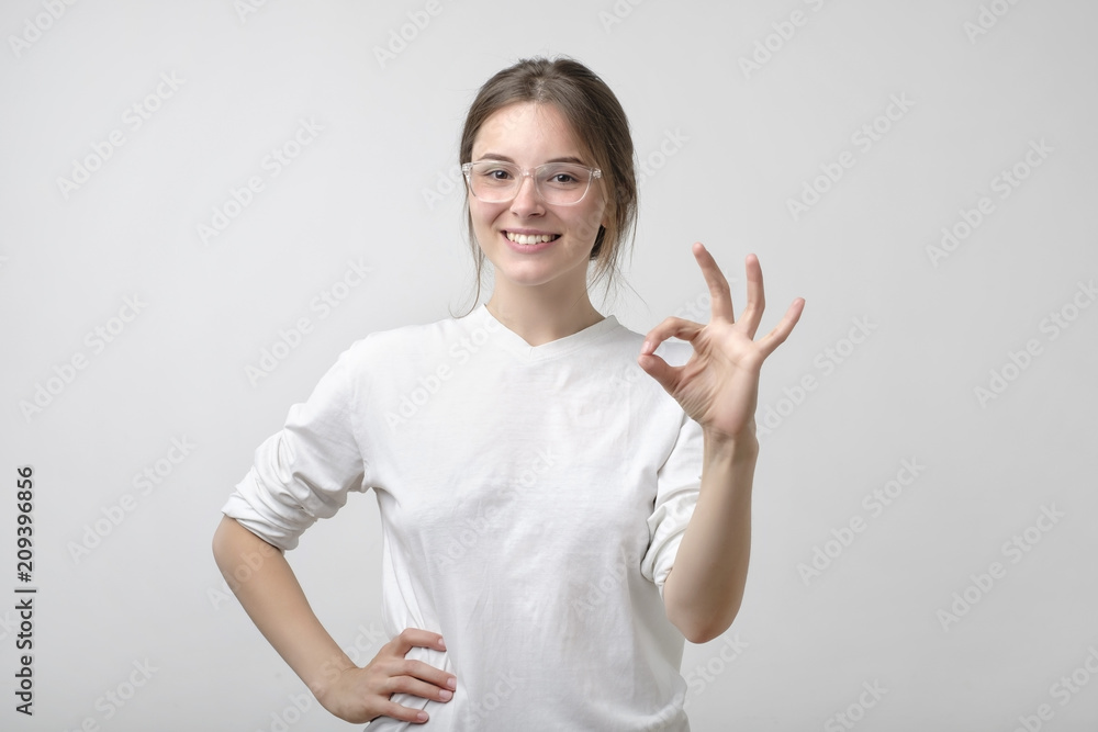Beautiful young woman in glasses is looking at camera, showing Ok sign and smiling.