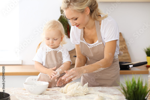 Little girl and her blonde mom in beige aprons  playing and laughing while kneading the dough in the kitchen. Homemade pastry for bread, pizza or bake cookies